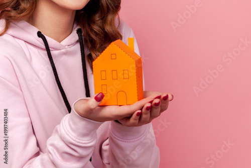 Cropped closeup view of unrecognizable woman holding paper house model representing real estate investment property planning and mortgage isolated on pink background with copy space