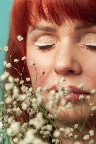 Close up of redhead woman enjoying floral scent of flowers for aesthetic skincare and professional beauty makeover isolated on turquoise background