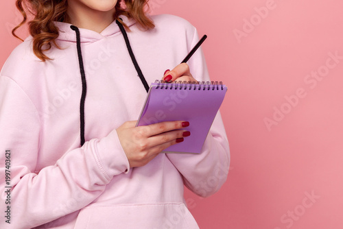 Cropped closeup view of woman writing in purple notebook with pencil writing isolated on pink background with copy space