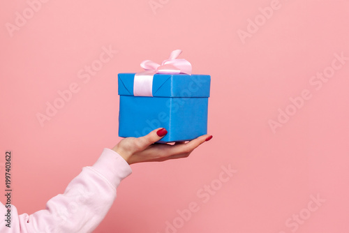 Cropped closeup view of anonymous woman's hands holding blue gift box with ribbon present in little parcel isolated on pink background with copy space