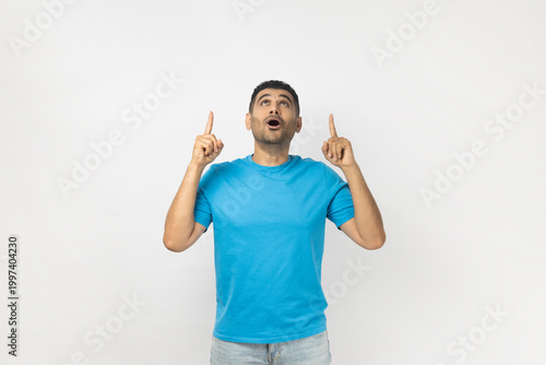 Portrait of amazed unshaven man wearing blue T- shirt standing pointing upwards with index fingers, looking up with big eyes and open mouth. Indoor studio shot isolated on gray background.