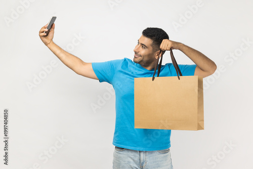 Portrait of attractive smiling positive unshaven man blogger wearing blue T- shirt standing with shopping bag, making selfie for his blog in mall. Indoor studio shot isolated on gray background.