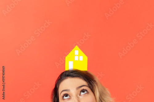 Anonymous person holding little paper house on head dreaming about apartment or planning to invest into building to ensure future housing security isolated over orange background with copy space