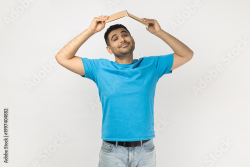 Portrait of clever smart good looking optimistic unshaven man wearing blue T- shirt standing holding book above head as a roof. Indoor studio shot isolated on gray background.