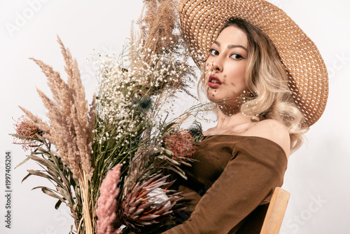 Pretty lady in straw hat posing with floral arrangement of pampas grass and protea looking through flowers at camera with shadow patterns on face skin isolated on white background