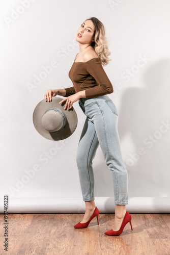 Attractive young woman holding wide brim hat while posing in profile wearing brown off-shoulder top blue jeans and red shoes looking at camera standing on wooden floor near white wall background