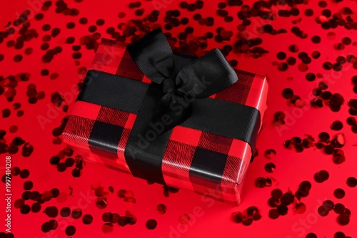 Gift Box Wrapped in Plaid Pattern Sits on Red Background With Scattered Confe...