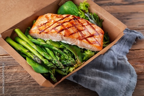 Grilled Salmon With Greens and Asparagus in a Takeout Container on a Wooden T...
