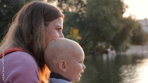 Mother holding her baby boy in sling carrier while he curiously points at something in park by water during sunset. Showing kid world around. Mom and baby walk together. Cognitive emotions of child