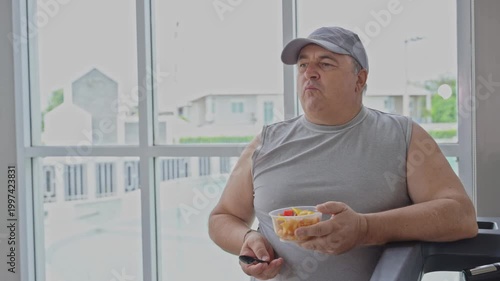 Overweight mature man eating healthy fresh fruit salad after workout near window