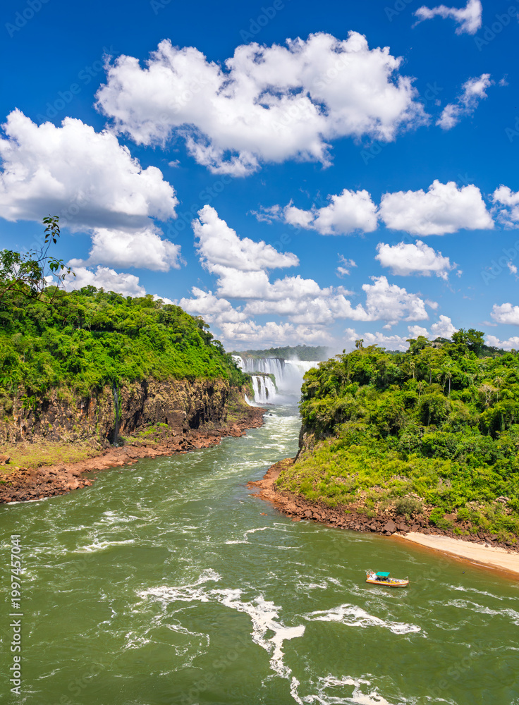 Naklejka premium Breathtaking panoramic view of the majestic Iguazu Falls with the river flowing through a lush green canyon and a tourist boat under a clear blue sky with fluffy white clouds