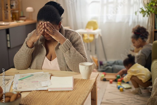 African American mother covering face while reviewing household bills, struggling with financial stress as children playing nearby. Ideal for poverty, debt, budgeting, family hardship themes