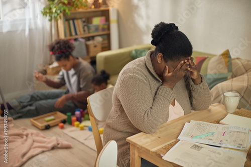 Фотография African American mother covering face while sitting by unpaid bills, children playing behind her at home