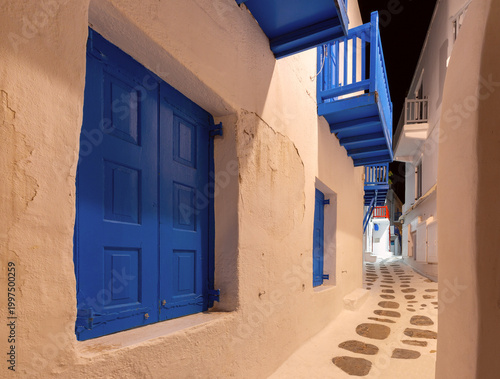 Night view of a narrow street in Mykonos, Greece, with whitewashed walls, colorful balconies, stone paving and warm lights in the quiet old town.