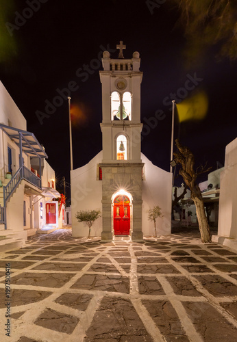 Night view of a traditional church in Mykonos, Greece, with an illuminated bell tower, red doors, stone paving and whitewashed walls in the quiet old town.
