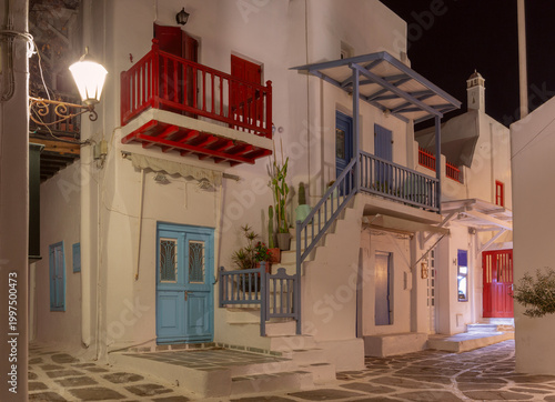 Night view of a narrow street in Mykonos, Greece, with whitewashed walls, colorful balconies, stone paving and warm lights in the quiet old town.