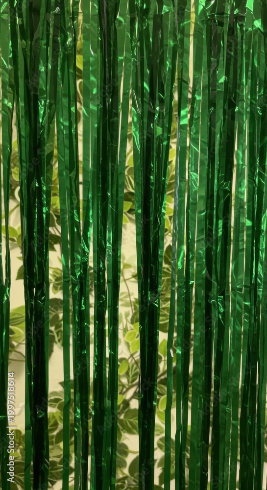 Fototapeta premium Green metallic foil fringe curtain hanging against a lush leaf wall background for party decorations, festive event styling and holiday celebratory backdrops