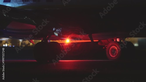 Low angle view of large aircraft underside at night, ground vehicle with yellow green marking visible beneath fuselage, runway light glowing in distance
