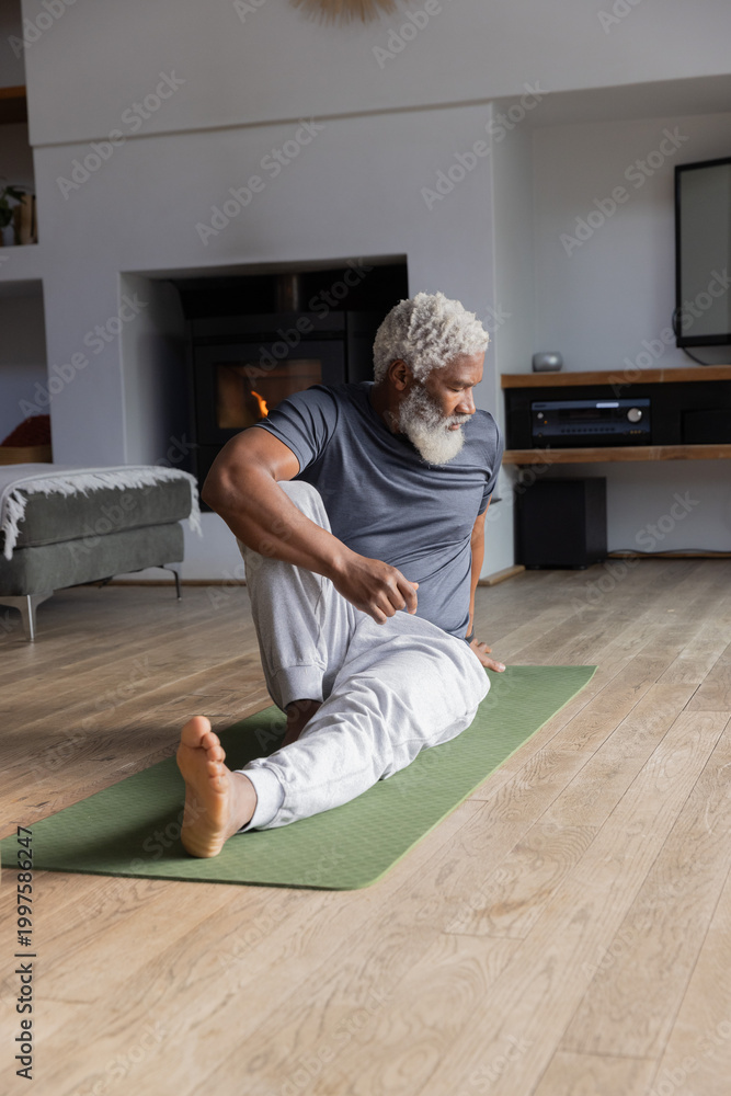 Naklejka premium Senior African American man performing seated twist on green mat in living room, wearing sweatpants