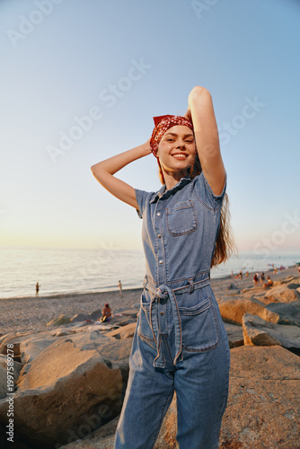 Lifestyle woman in modern boho-western denim street style smiling confidently outdoors near rocky beach with warm film color tone at sunset.