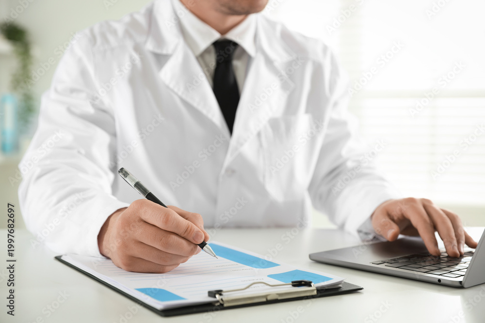 Fototapeta premium Doctor in medical coat working with clipboard and laptop at white table in clinic, closeup