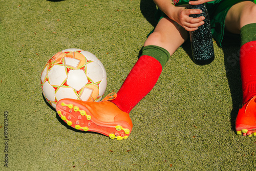 Young girl in red socks and orange soccer cleats sits on green grass next to a white soccer ball, holding a black water bottle in a sunny outdoor setting