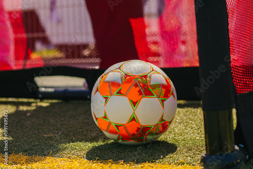 Football with vibrant orange and white design resting on artificial turf near goalpost, with netting and blurred players in the background during a girls' football match