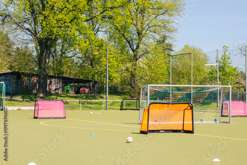 Girls' football training field with small goals and soccer balls scattered across the artificial turf, surrounded by trees and a storage shed in the background