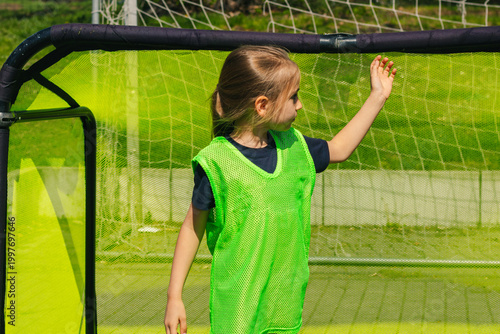 Young girl wearing a bright green training vest stands near a soccer goal on a sunny day, preparing for a girls' football practice session in an outdoor field
