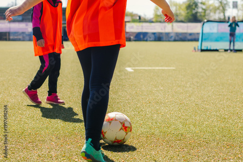 Girls' football players in bright orange jerseys dribble a soccer ball on a green artificial turf field with training goals visible in the background