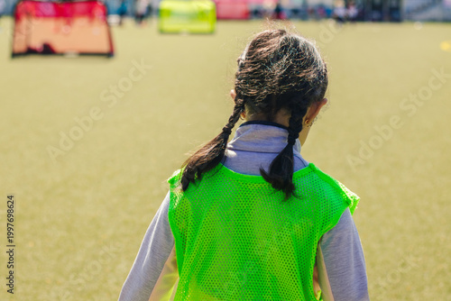 Young girl wearing a bright green training vest stands on a football field, with small goal nets visible in the background under clear blue skies