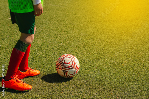 Young girl in green sportswear and red socks stands beside a soccer ball on an artificial turf field, sunlight illuminating the scene from the right side