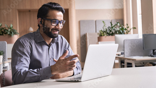 Happy indian business man remote teacher, customer support manager wearing headset talking at virtual meeting consulting client on video call giving distance learning class at home office call center.
