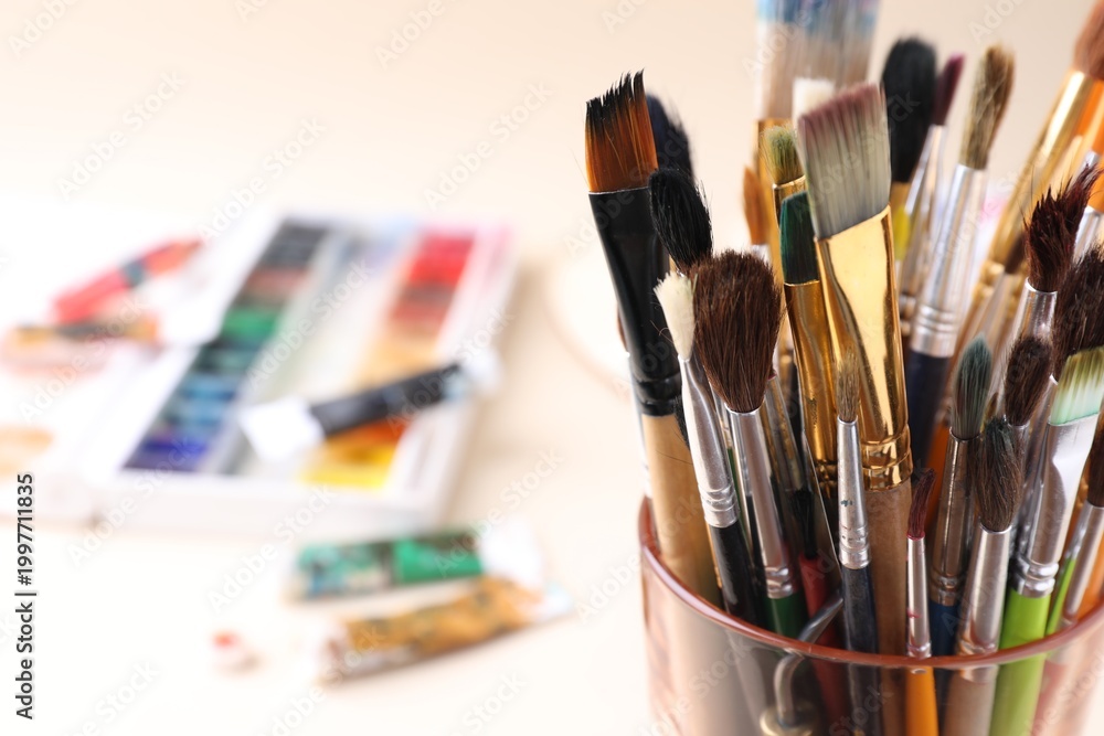 Fototapeta premium Paintbrushes in glass and paints on beige background, selective focus. Space for text