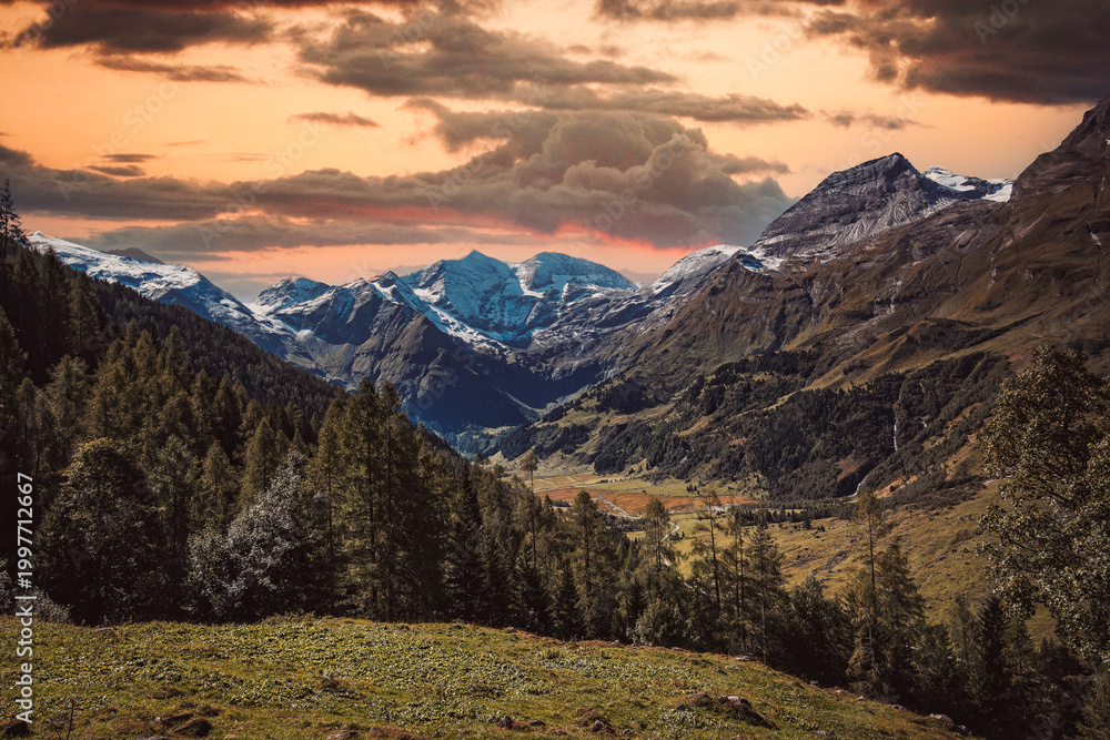 Fototapeta premium Peaceful picturesque mesmerizing atmospheric landscape with high Snow-capped mountains, Großglockner alpine serpentine pass road, and meadow grasslands pasture in sunset