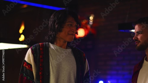 Two male friends laughing and talking together while one holds an electronic cigarette in a nightclub with neon lights