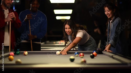 Cheerful young woman making a winning billiards shot while playing pool with multicultural friends in a dark bar
