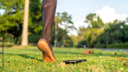 A dark-skinned foot in focus on a green lawn, near a phone and trees