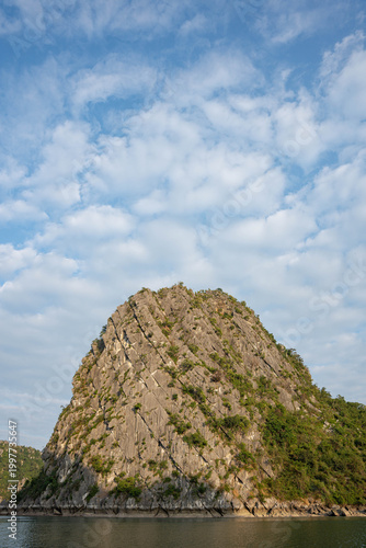 a dramatic limestone karst island rising steeply from calm waters under a bright, cloud-filled sky