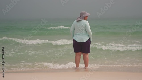 Slow Motion Plus size woman in her 50s stands at the surf line as the sun breaks through a marine layer of mist and fog