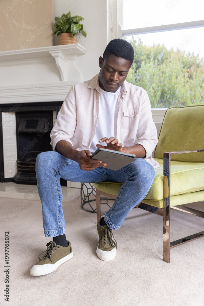 Naklejka premium African American man sitting in living room on green armchair using tablet and tapping screen