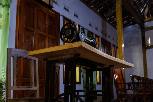 Vintage ornate black and gold treadle sewing machine on a white table in a rustic room with green curtains and wooden door. Classic antique craftsmanship and traditional interior.