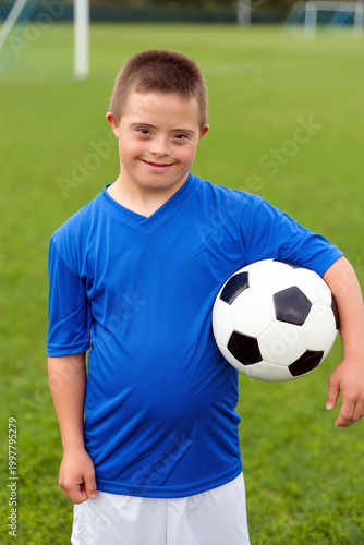 A cheerful child with Down syndrome stands on a lush green soccer field, holding a black and white soccer ball, ready for practice. Generative AI.