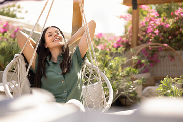 Woman leans back in cozy swing chair with hands behind head, enjoying comfortable outdoor home furniture and peaceful leisure, surrounded by plants and flowers on sunny terrace. Wellbeing, relaxation
