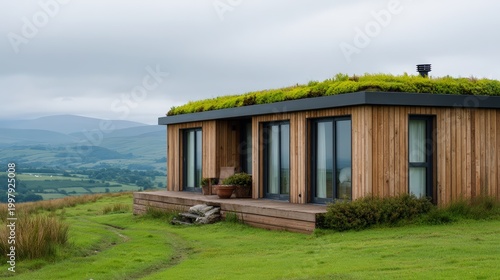 A modern eco-friendly wooden house with large windows and a green roof sits amidst lush rolling hills under a cloudy sky.