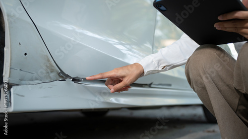 Fotografía Close-up of a car’s lower side panel with visible dent and paint damage as a person points to the scrape while holding a clipboard, suggesting vehicle inspection or insurance claim