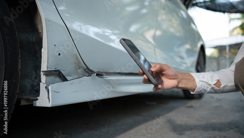 Close-up of damaged car rocker panel near wheel while a person photographs the dent with a smartphone for insurance claim or repair estimate outdoors.