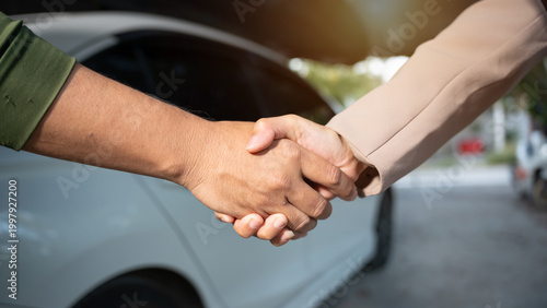 Close-up handshake between two people next to a white car, suggesting agreement during vehicle sale or service transaction, outdoor daylight, shallow depth of field.