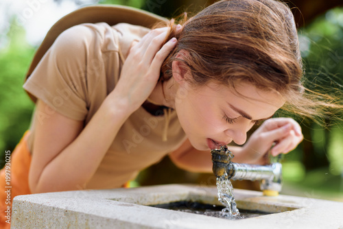 Woman drinking water from a concrete fountain in an outdoor park during summer, young female hydrating in casual clothes, closeup profile showing refreshment and natural sunlight.