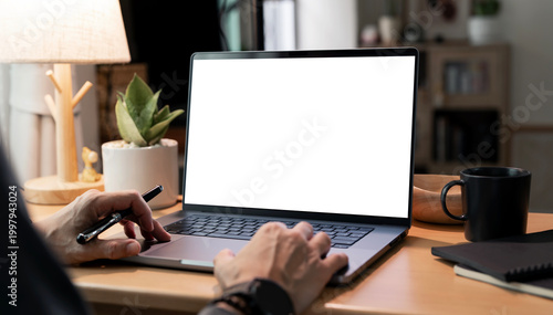 Laptop mockup on wooden desk with blank white screen, hands typing, cozy home office workspace.